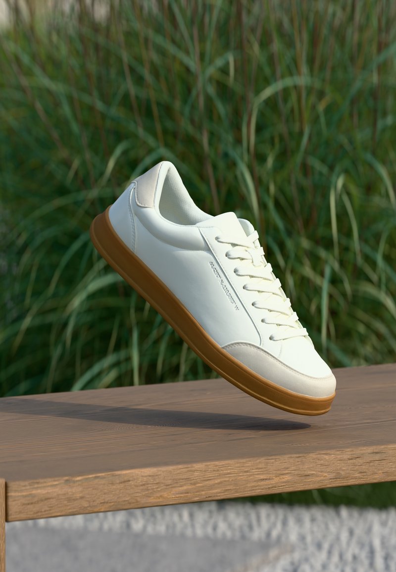 Baskets blanches avec des accents beiges et une semelle en gomme flottant au-dessus d'un banc en bois, avec un fond d'herbe verte sous une lumière douce du jour.