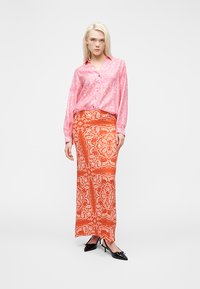 Γυναίκα που στέκεται, φορώντας μια ροζ πουκάμισο με κουμπιά και μοτίβο, και μία πορτοκαλί μακριά φούστα με λευκά σχέδια πεϊσλί, συνδυασμένο με μαύρες μυτερές γόβες.