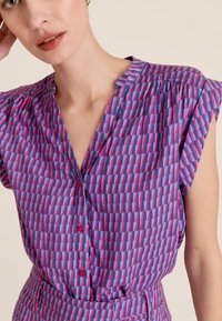 Blouse à manches courtes avec un col en V, présentant un motif géométrique en rose et bleu sur un tissu texturé, rehaussée de boutons roses.