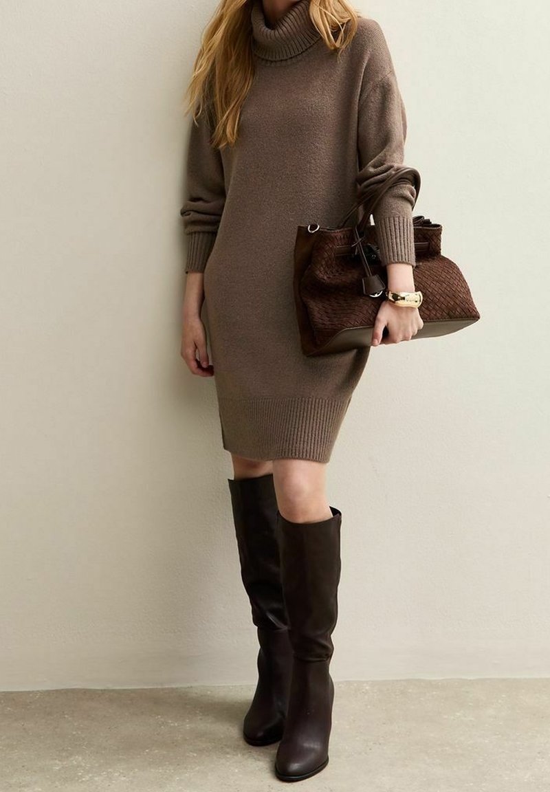 Femme portant une robe-pull marron à col roulé, tenant un grand sac à main tressé marron foncé, et portant des bottes noires montant jusqu'aux genoux.