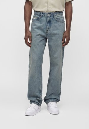 Mand iført lyseblå jeans med lige ben, hvide sneakers og en beige kortærmet skjorte stående på en ensfarvet baggrund.