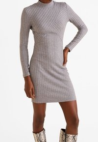 Robe grise côtelée avec un col haut et des manches longues, ornée de détails de couture diagonale. Le modèle porte des bottes métalliques imprimées serpent.