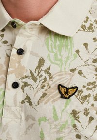 Polo shirt con tessuto testurizzato beige caratterizzato da motivi floreali verdi, bottoni neri e un'etichetta con logo ricamato dorato a forma di ali.