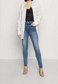 Bílá nadměrná button-up bunda přes černý saténový top, zkombinovaná s modrými skinny džíny a béžovými hadovinovými lodičkami.