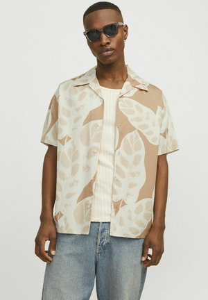 Chemise à manches courtes en tissu beige et crème, arborant un grand motif de feuilles. Boutons sur le devant, associée à un débardeur côtelé en dessous.