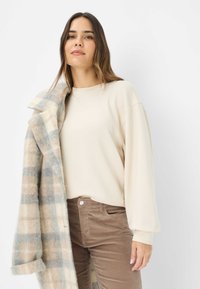 Crème-kleurige sweatshirt met lange mouwen, gecombineerd met bruine ribfluwelen broek. Een geruite wollen jas in beige en blauwe tinten hangt over de schouder.