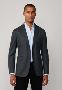 Dunkelgrauer Blazer aus strukturiertem Stoff, mit zwei Vordertaschen und einem einzigen Knopfverschluss, kombiniert mit einem hellblauen Hemd darunter.