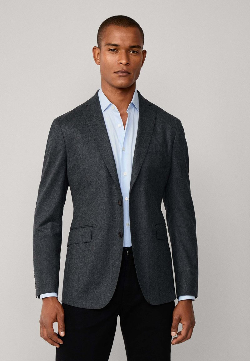 Dunkelgrauer Blazer aus strukturiertem Stoff, mit zwei Vordertaschen und einem einzigen Knopfverschluss, kombiniert mit einem hellblauen Hemd darunter.