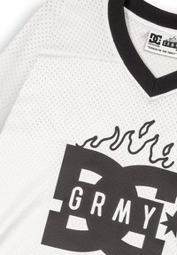 Biała sportowa koszulka z siateczki z czarnym dekoltem w kształcie V, z dużym czarnym logo "DC" z płomieniami i literami "GRMY" na piersi.