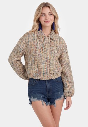 Frau mit blondem Haar, die eine mehrfarbige, texturierte Langarmjacke, blaue Quastenohrringe und hoch taillierte, ausgefranste Jeansshorts trägt.