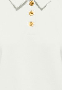 Chemise blanche à col avec trois boutons dorés en forme de soleil alignés sur le devant.