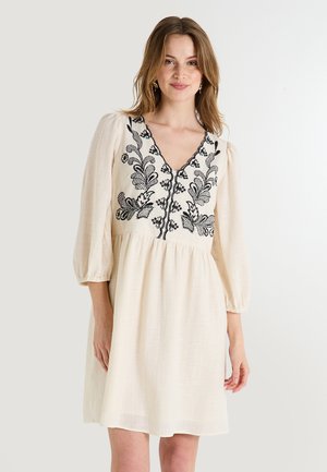 Robe de couleur crème avec un décolleté en V, des manches bouffantes et une broderie florale noire complexe sur le corsage et les boutons. Tissu texturé.