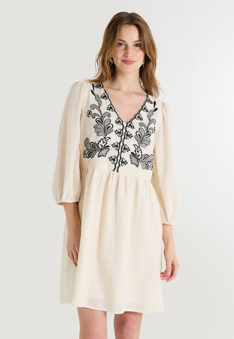 Vestido color crema con escote en V, mangas abullonadas y delicada bordadura floral negra a lo largo del cuerpo y los botones. Tela texturizada.