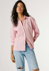 Ženska z dolgimi kodrastimi lasmi, oblečena v ohlapno roza srajco z gumbi in svetlo modre strgane kavbojke, stoji pred enobarvnim svetlim ozadjem.