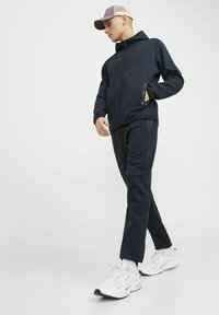 Haut à capuche navy avec fermeture éclair et poches, associé à un pantalon assorti. Porté avec des chaussures de sport blanches et une casquette rose clair.