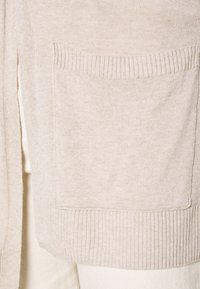 Béžový pletený kardigan s žebrovaným lemem a detailem kapsy. Textura je měkká, s hladkým a rovnoměrným vzorem po celém materiálu.