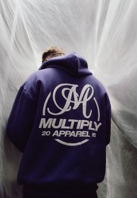 Lila Kapuzenpullover mit einem großen weißen Grafiklogo und dem Text "MULTIPLY 20 APPAREL 15" auf dem Rücken, mit einer Zugkordel-Kapuze.