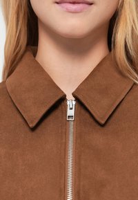 Καφέ σακάκι από σουέντ με γιακά, που διαθέτει ασημένιο φερμουάρ και ομαλή υφή. Σημαντική λεπτομέρεια είναι ο καθαρός, μινιμαλιστικός σχεδιασμός.