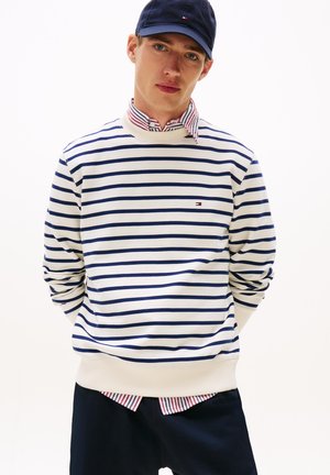 Gestreifter Pullover in Creme und Marineblau, mit gerippten Bündchen und Saum, über einem rot-weißen karierten Hemd getragen, dazu eine marineblaue Mütze.