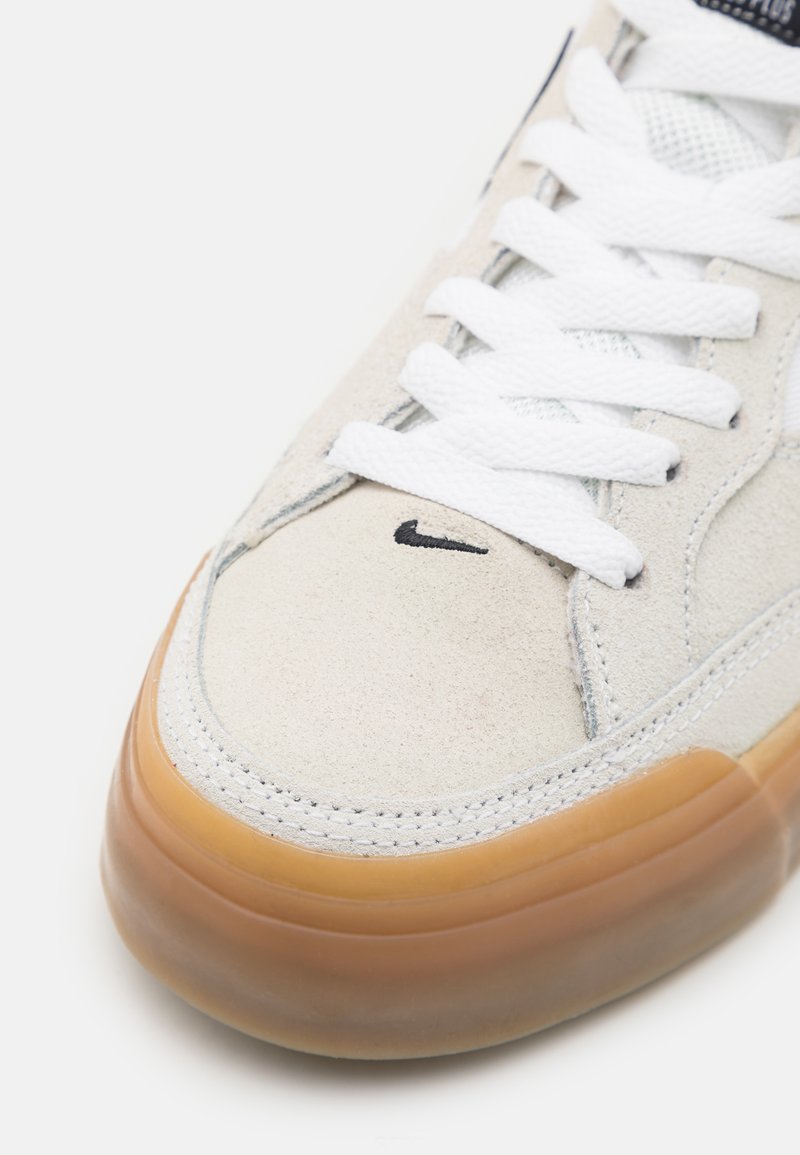 Zapatilla Nike con parte superior de ante gris claro, cordones blancos, un logo negro en la puntera y una suela transparente de goma.