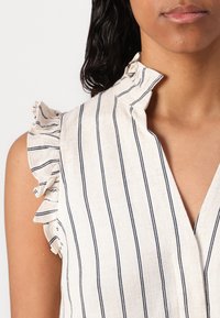 Blouse sans manches à rayures avec un col en V, dotée de bords volantés aux épaules. Le tissu est d'un beige léger et texturé avec des rayures bleu foncé.