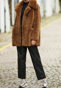 Abrigo marrón esponjoso con cuello ancho y bolsillos parche, combinado con pantalones de pierna ancha de cuero sintético negro y zapatillas metálicas. Diseño texturizado y de gran tamaño.