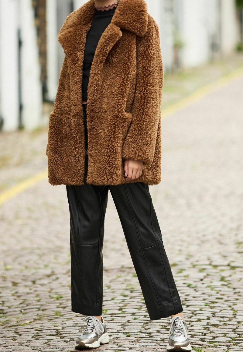 Abrigo marrón esponjoso con cuello ancho y bolsillos parche, combinado con pantalones de pierna ancha de cuero sintético negro y zapatillas metálicas. Diseño texturizado y de gran tamaño.