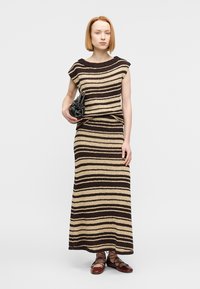 Žena stojaca, oblečená v bezrukávovom hnedo-bežovom pruhovanom pletenom topu a dlhom sukni v rovnakom vzore, drží čiernu kabelku a má na sebe hnedé šnurovacie topánky.