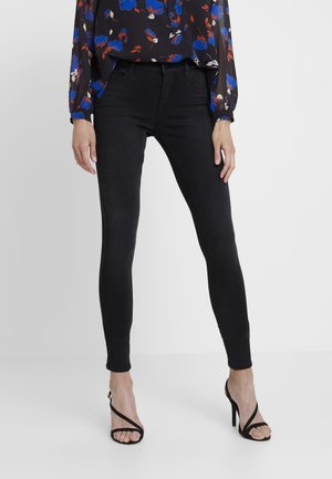 Femme portant un jean skinny noir, des talons hauts noirs à lanières, et un chemisier noir à imprimé floral bleu et rouge, debout devant un fond blanc.