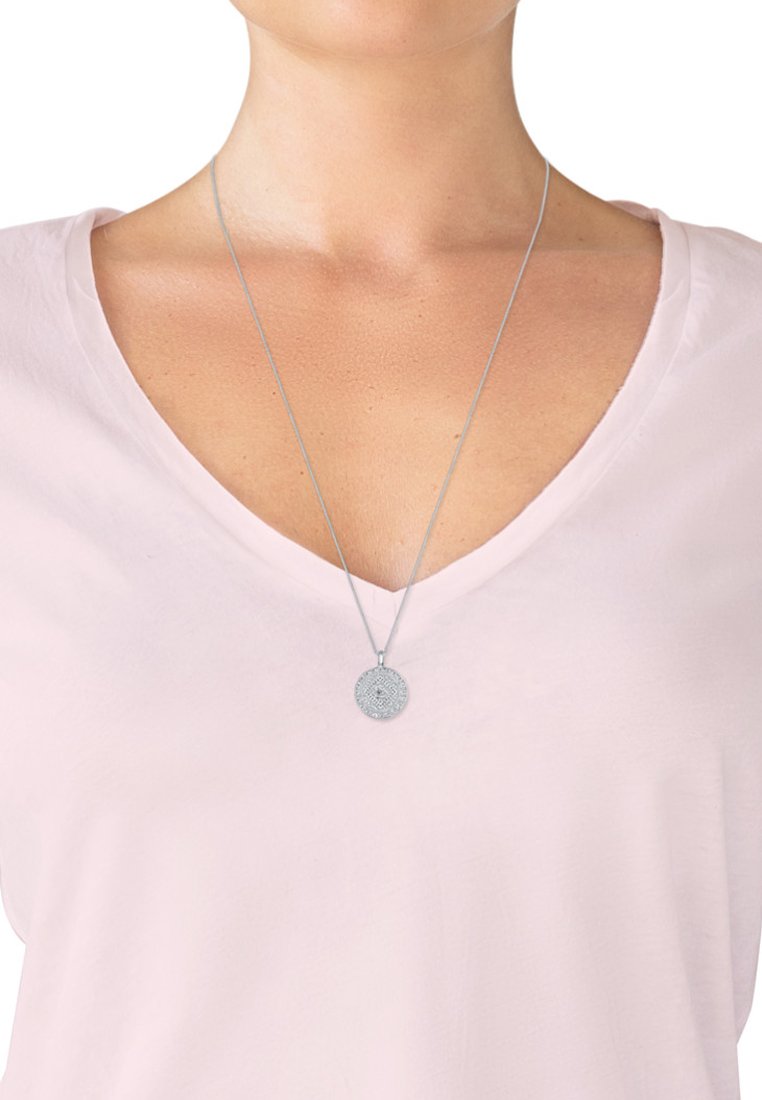 Collana con pendente in argento dal design circolare e intricato, appesa a una sottile catena, indossata sopra una maglietta a V rosa chiaro.