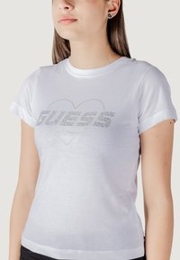 Witte katoenen t-shirt met korte mouwen en een aansluitend ontwerp. De voorkant heeft een zilveren strass 'GUESS' logo met een hartomtrek.