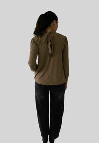 Vrouw met donker haar in een paardenstaart, gekleed in een bruine lange mouwtop met een strik aan de achterkant en zwarte jeans, staand tegen een effen grijze achtergrond.