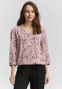 Femeie cu păr lung, castaniu, purtând o bluză roz cu model, cu decolteu în V și mâneci trei sferturi, împreună cu pantaloni negri, stând în fața unui fundal simplu.