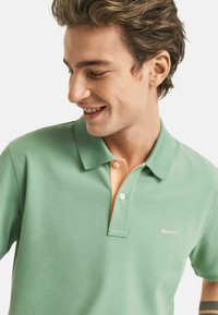 Polo shirt verde con una morbida texture, caratterizzato da un placket in un contrastante arancione chiaro e due bottoni. Piccolo logo GANT in oro sul petto.