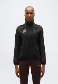 Γυναίκα που φοράει μαύρο αθλητικό μπουφάν με φερμουάρ, με τα λογότυπα "World Series of Fitness Racing" και "Hyrox", συνδυασμένο με σκουρό καφέ κολάν.