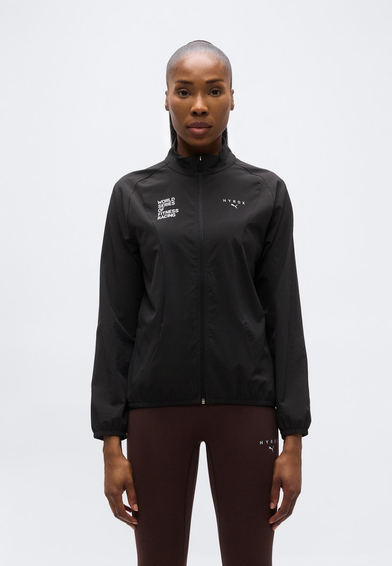 Γυναίκα που φοράει μαύρο αθλητικό μπουφάν με φερμουάρ, με τα λογότυπα "World Series of Fitness Racing" και "Hyrox", συνδυασμένο με σκουρό καφέ κολάν.
