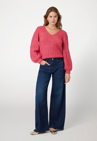 Maglione lavorato a maglia rosa con scollo a V e texture a costine, abbinato a jeans in denim blu scuro a gamba larga e ballerine in oro lucido.