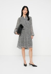 Γυναίκα φορώντας ένα μαύρο και άσπρο μοτίβο φόρεμα έως το γόνατο, κρατώντας μια μεγάλη μαύρη τσάντα, και φοράει μαύρα χαμηλοτάκουνα παπούτσια.