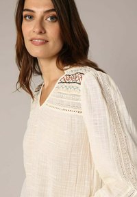 Witte textuur blouse met ingewikkelde borduursels op de schouders, kantdetails langs de mouwen en een v-hals ontwerp.