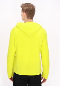 Giacca a vento gialla brillante, realizzata in materiale leggero, con maniche lunghe e una texture liscia, caratterizzata da un design sportivo.