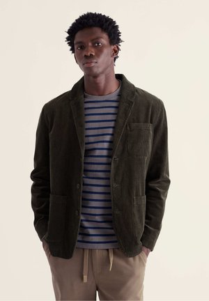 Jeune homme portant un blazer en velours côtelé vert foncé sur une chemise rayée grise et marine avec un pantalon beige, debout les mains dans les poches.