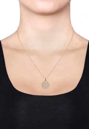 Rund, rosaguld vedhængs halskæde med et geometrisk blomstermønster, hængende på en tynd kæde, båret mod en sort bluse.