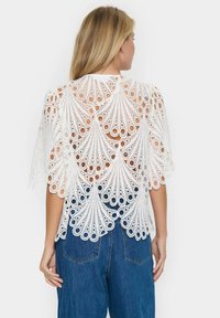 Top bianco in pizzo con motivi intagliati intricati e maniche corte, abbinato a jeans blu a vita alta.