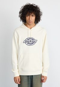 Kremfarget hettegenser med frontlomme og ribbede mansjetter. Har en blå logo med et sirkulært tau-design og teksten "Dickies."