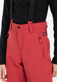 Pantaloni da sci rossi con bretelle nere regolabili, dotati di una tasca laterale con zip e un logo nero. Il tessuto sembra liscio e resistente.