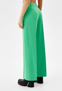 Kvinna som bär höga, vida gröna byxor, en kort brun topp och svarta plattformsklackar, stående mot en vit bakgrund.