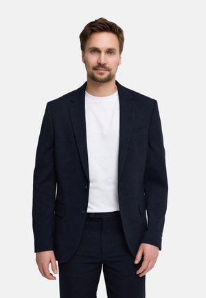 Mężczyzna o jasnobrązowych włosach i brodzie, ubrany w granatową marynarkę i spodnie na białej koszulce z okrągłym dekoltem, stojący na jednolitym tle.