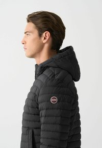 Chaqueta de abrigo negra con capucha, que presenta un diseño acolchado, textura suave y un parche de logo en el hombro izquierdo.