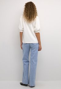 Witte korte mouwen top met pofmouwen, gecombineerd met lichtblauwe, uitlopende jeans. De outfit wordt aangevuld met zwarte open tenen schoenen.