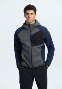 Giovane uomo con capelli ricci che indossa una giacca con cappuccio grigia e blu navy, e pantaloni neri, con le mani nelle tasche, rivolto in avanti.
